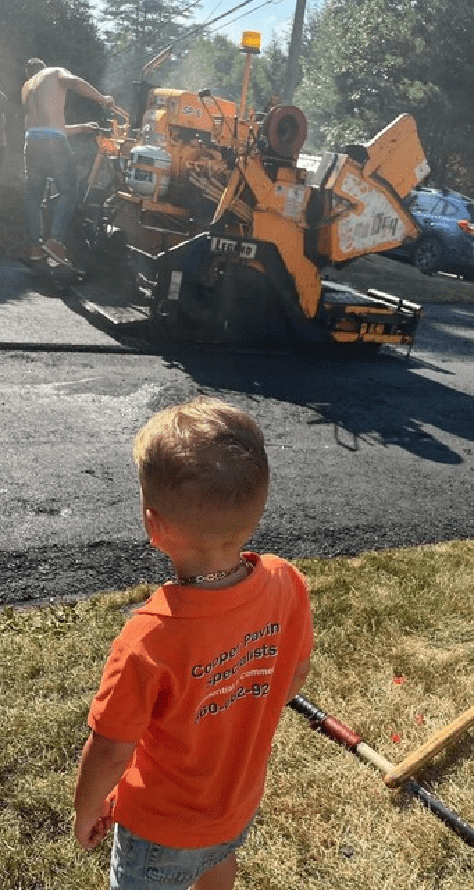 A child watching a Cooper Paving Specialists worker operating paving machinery, representing the next generation learning from the family-run paving company.